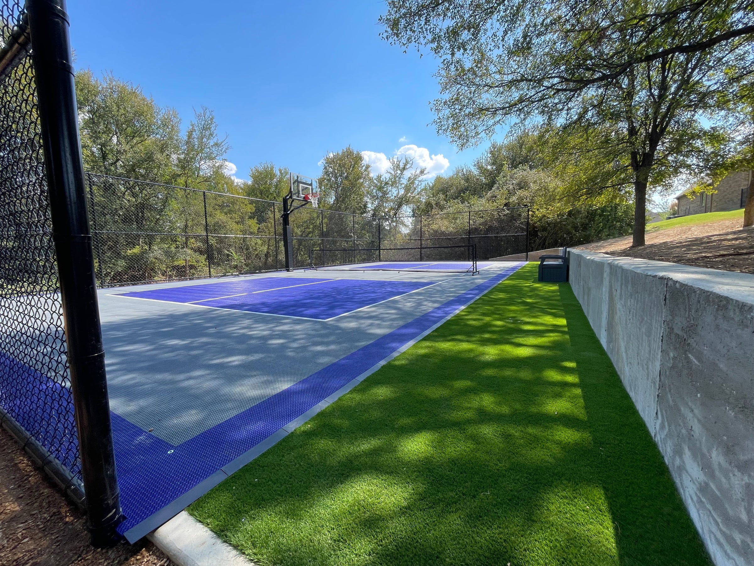 Backyard with a blue and white sports court, surrounded by trees and a clear sky.